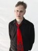 Roger on a plain white background wearing a black dress shirt with an open collar and a loose bright red necktie with a skeleton printed on the bottom